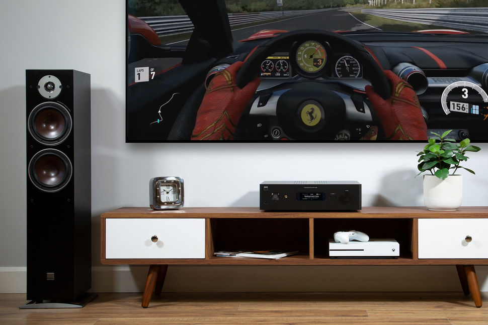 NAD C 399 BluOS Hybrid Digital DAC Streaming Amplifier on a TV bench with TV and DALI Oberon 7 speakers in the context of a recreation or living room or such. 